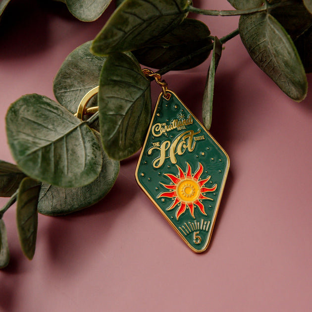 Decorative keychain with sun design and text, hanging among green leaves on a pink background